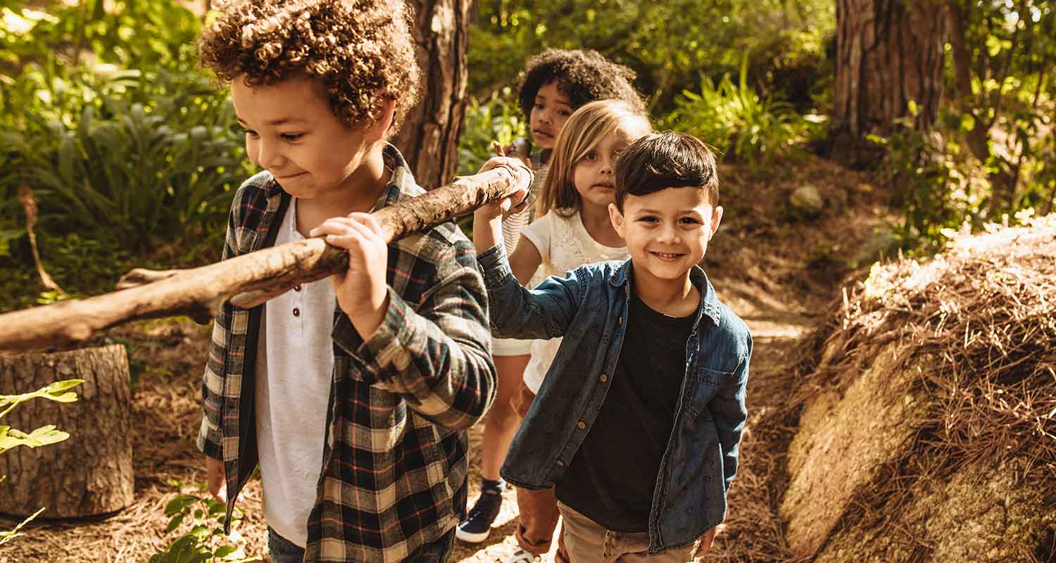 A group of 4 children smile whilst carrying a large wooden stick through the woods